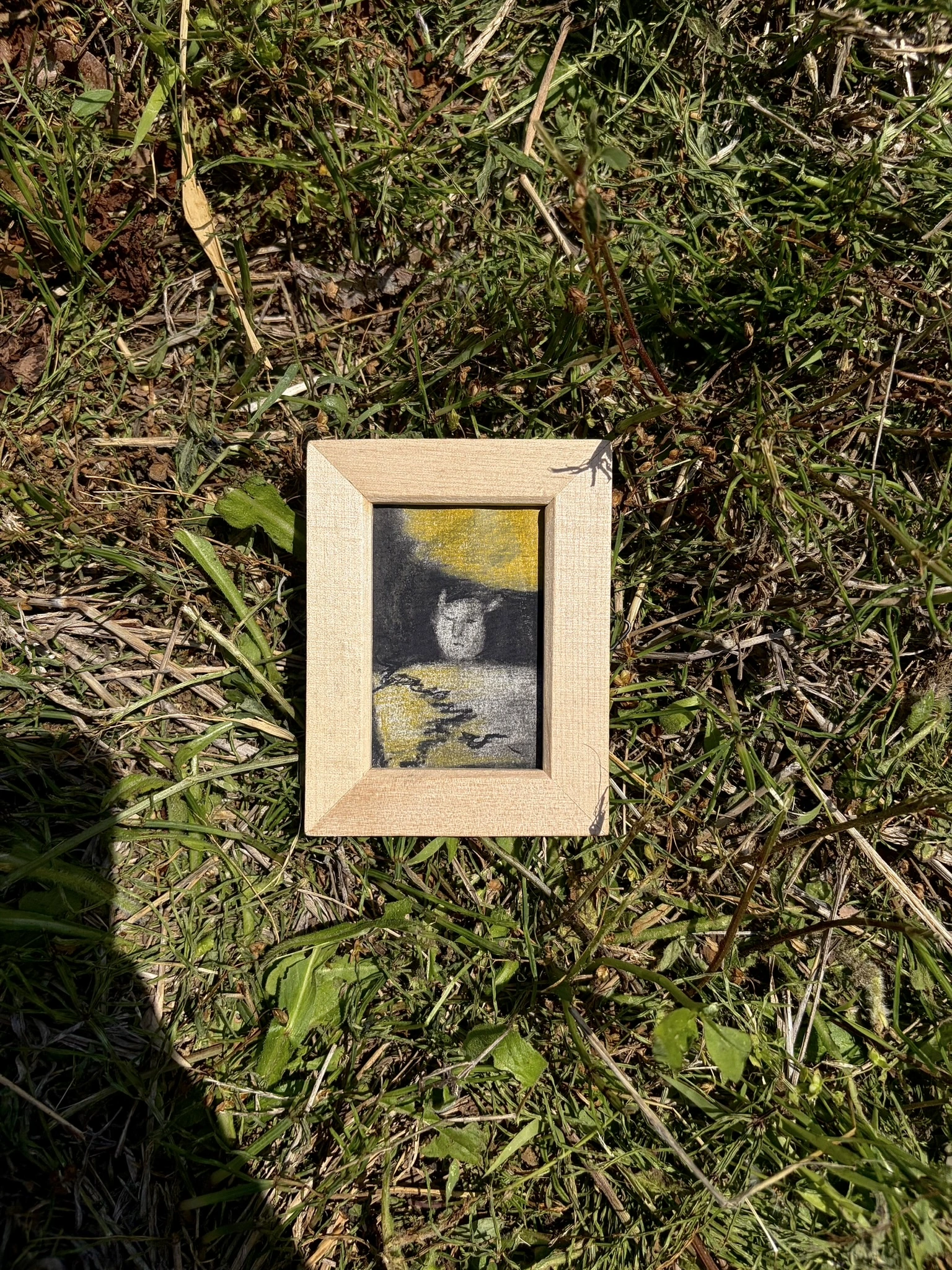 草の上に置いた木製の額に、人物の顔と黄色い風景を描いた小さな作品